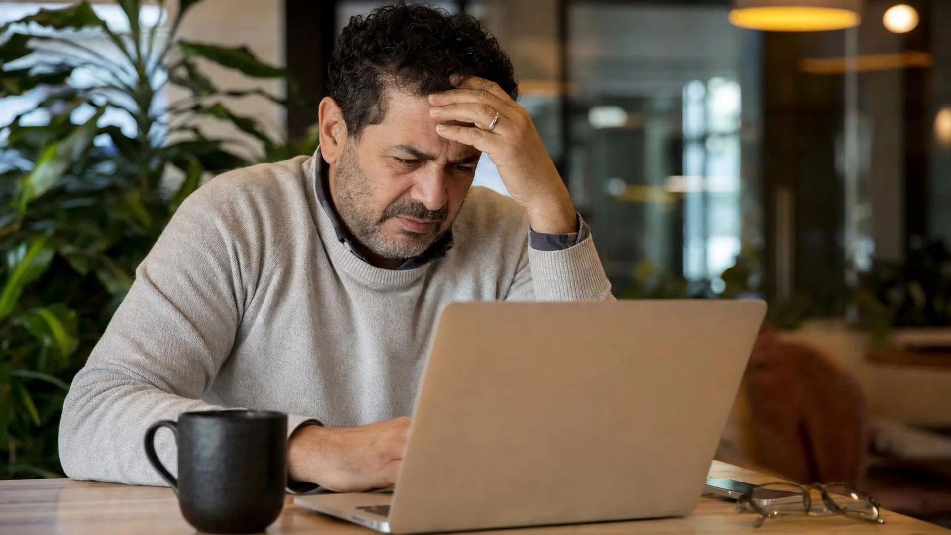 Persoon die met moeite naar een laptopscherm kijkt, hand op het voorhoofd, op een werkplek in een café