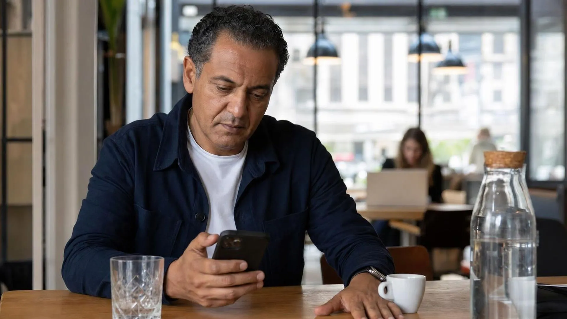 Persoon aan een cafétafel die met gespannen houding naar zijn telefoon staart