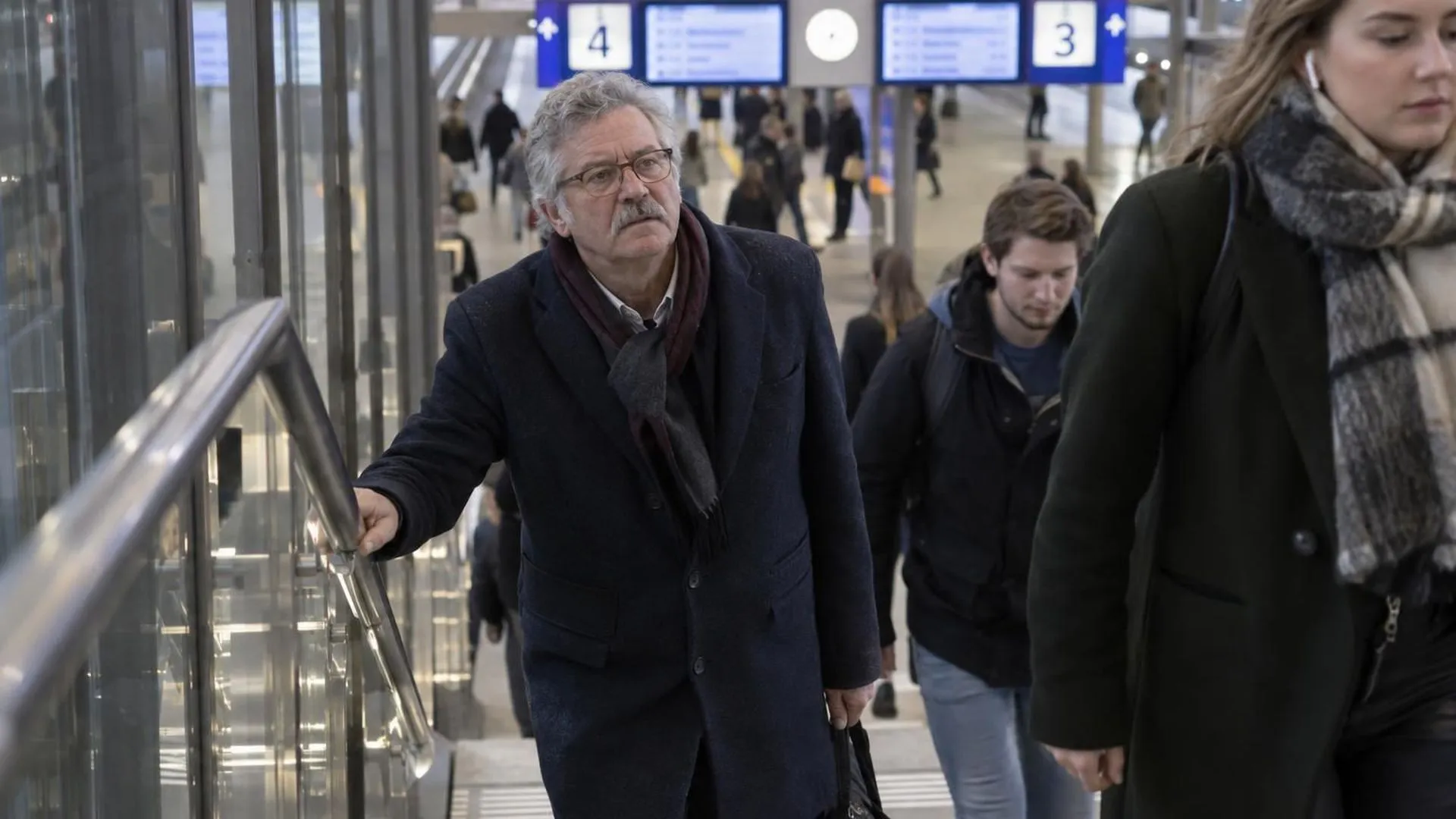 Iemand staat halverwege een stationstrap stil met een hand op de leuning, terwijl forenzen voorbij lopen