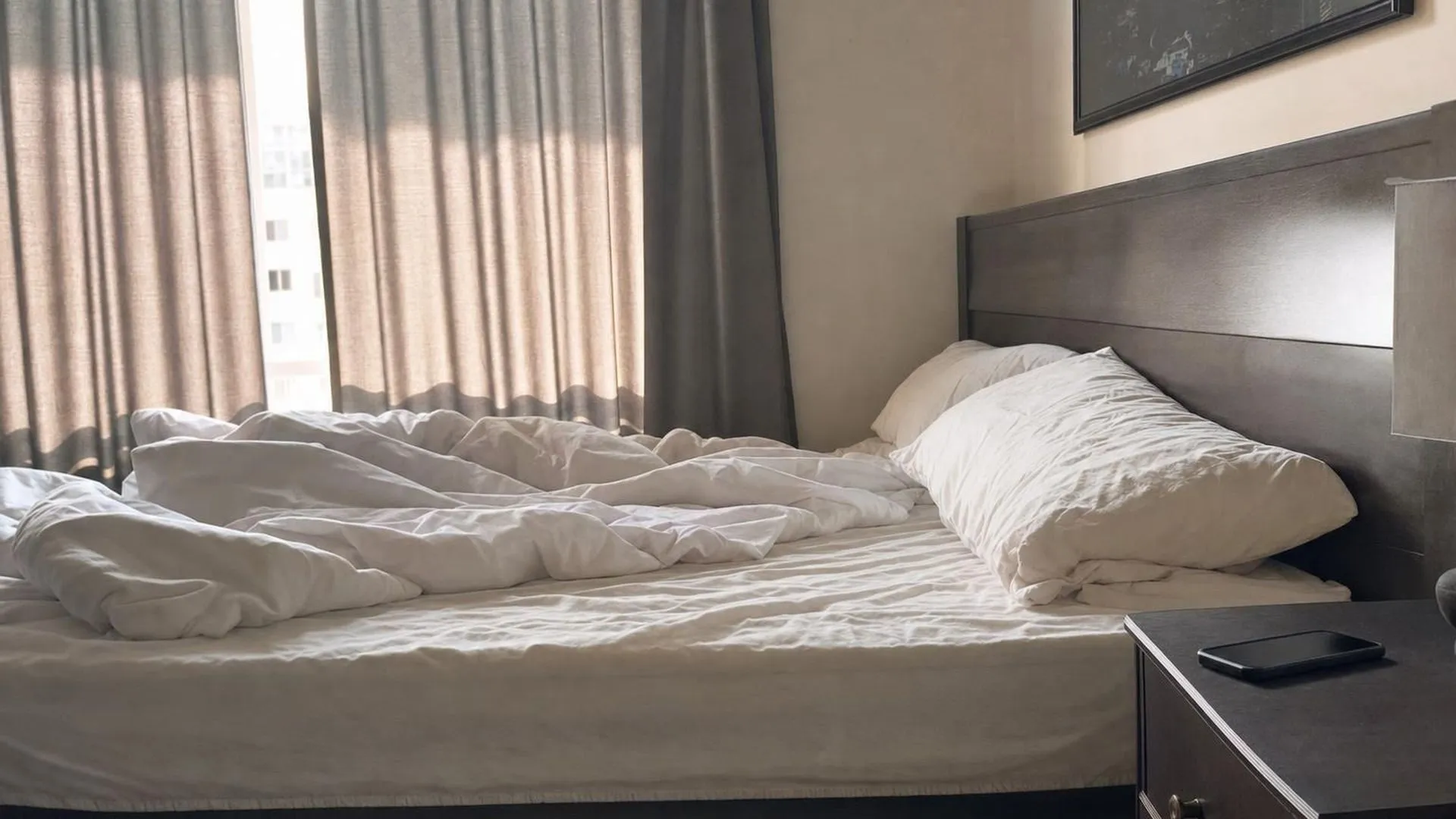 Onopgemaakt bed met daglicht door half dichte gordijnen en een telefoon op het nachtkastje