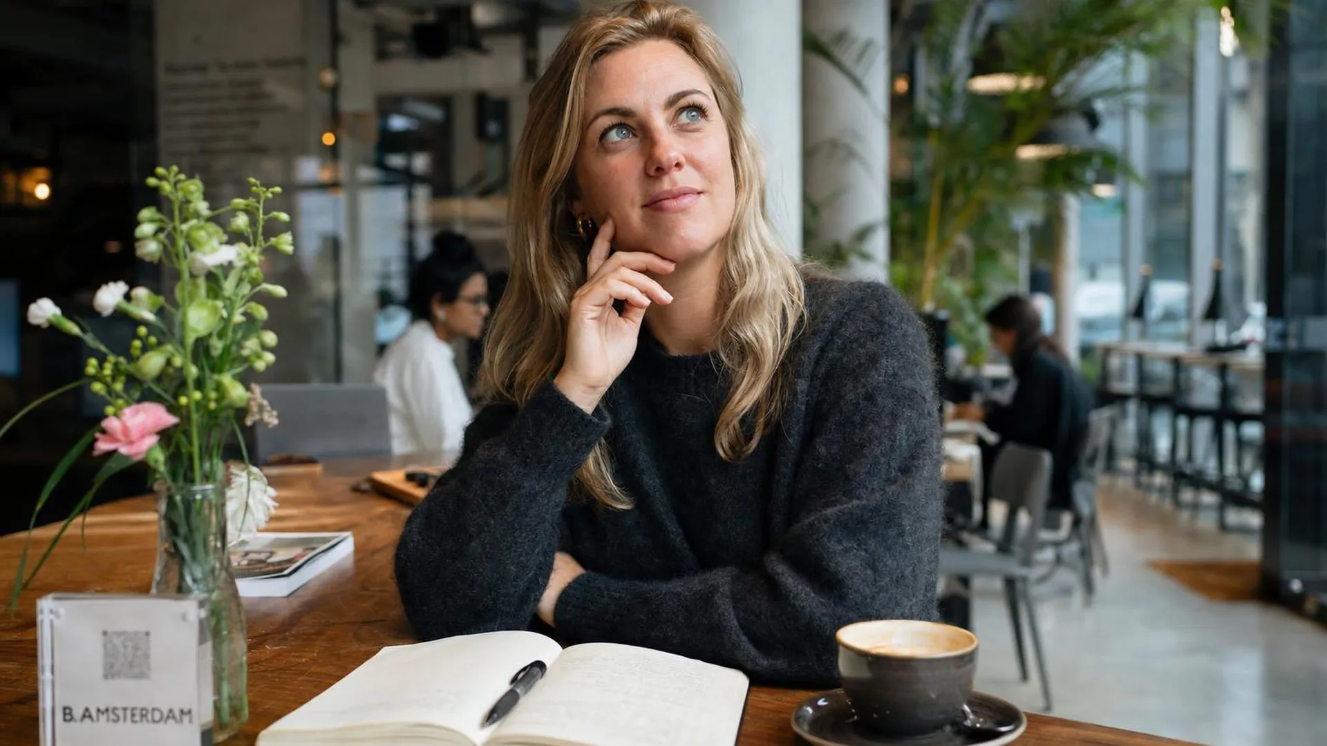 Persoon met notitieboek en koffie in een café, peinzend omhoog kijkend
