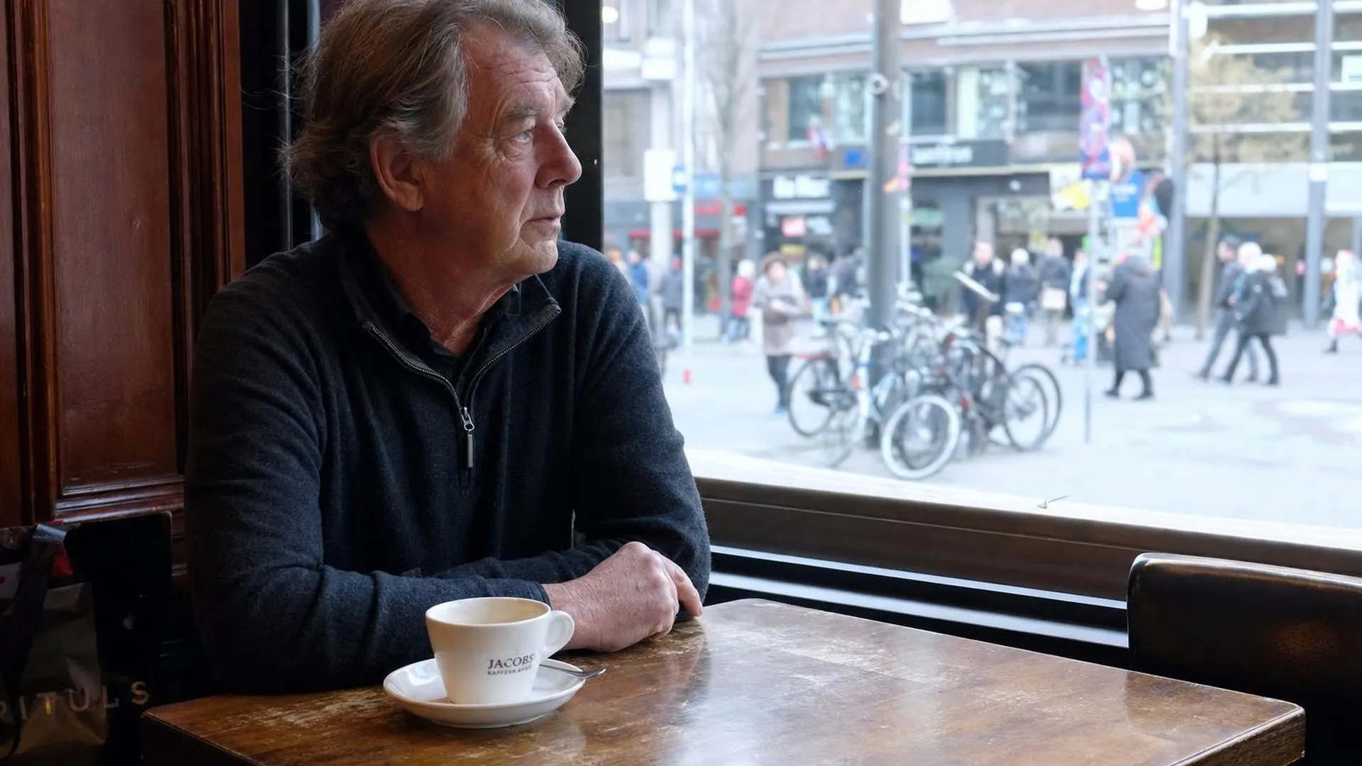 Iemand zit in gedachten aan een cafétafel, afwezig starend naar buiten