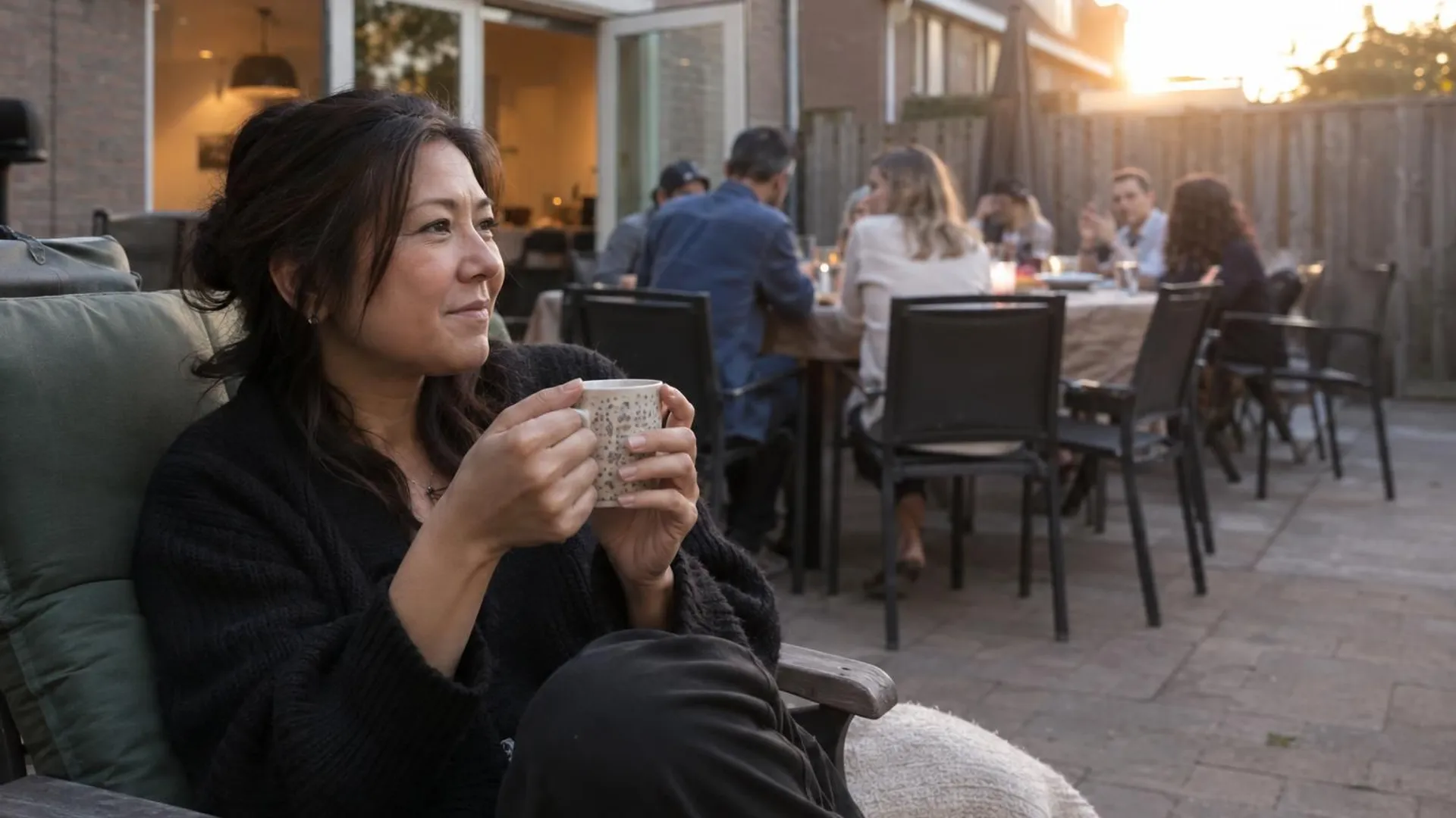 Een vrouw zit met een mok thee in een tuinstoel, iets apart van het gezelschap aan tafel in een achtertuin, in de avondzon.