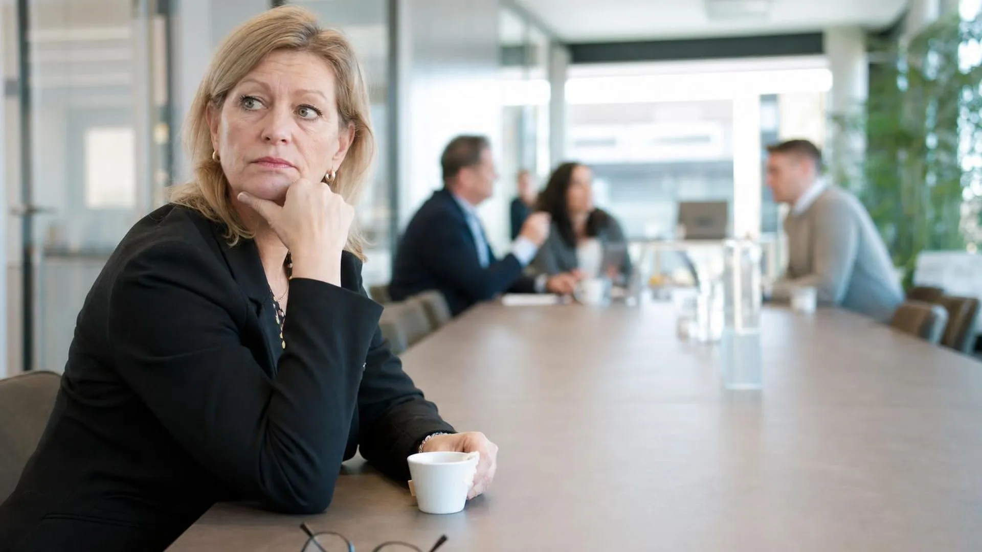Persoon zit alleen aan vergadertafel terwijl collega's op afstand staan