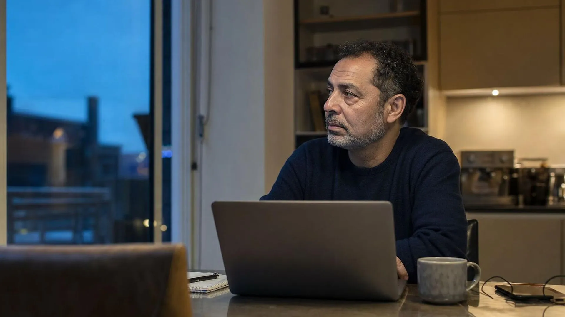 Persoon zit 's avonds aan de keukentafel met een open laptop en kijkt peinzend naar buiten